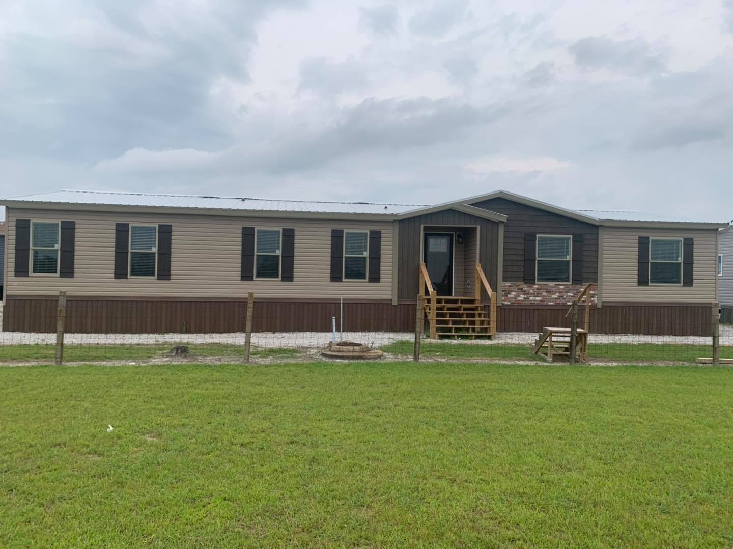 A beige and brown mobile home with black shutters and a small porch stands on a grassy lawn. The sky is overcast, suggesting a calm, subdued atmosphere.