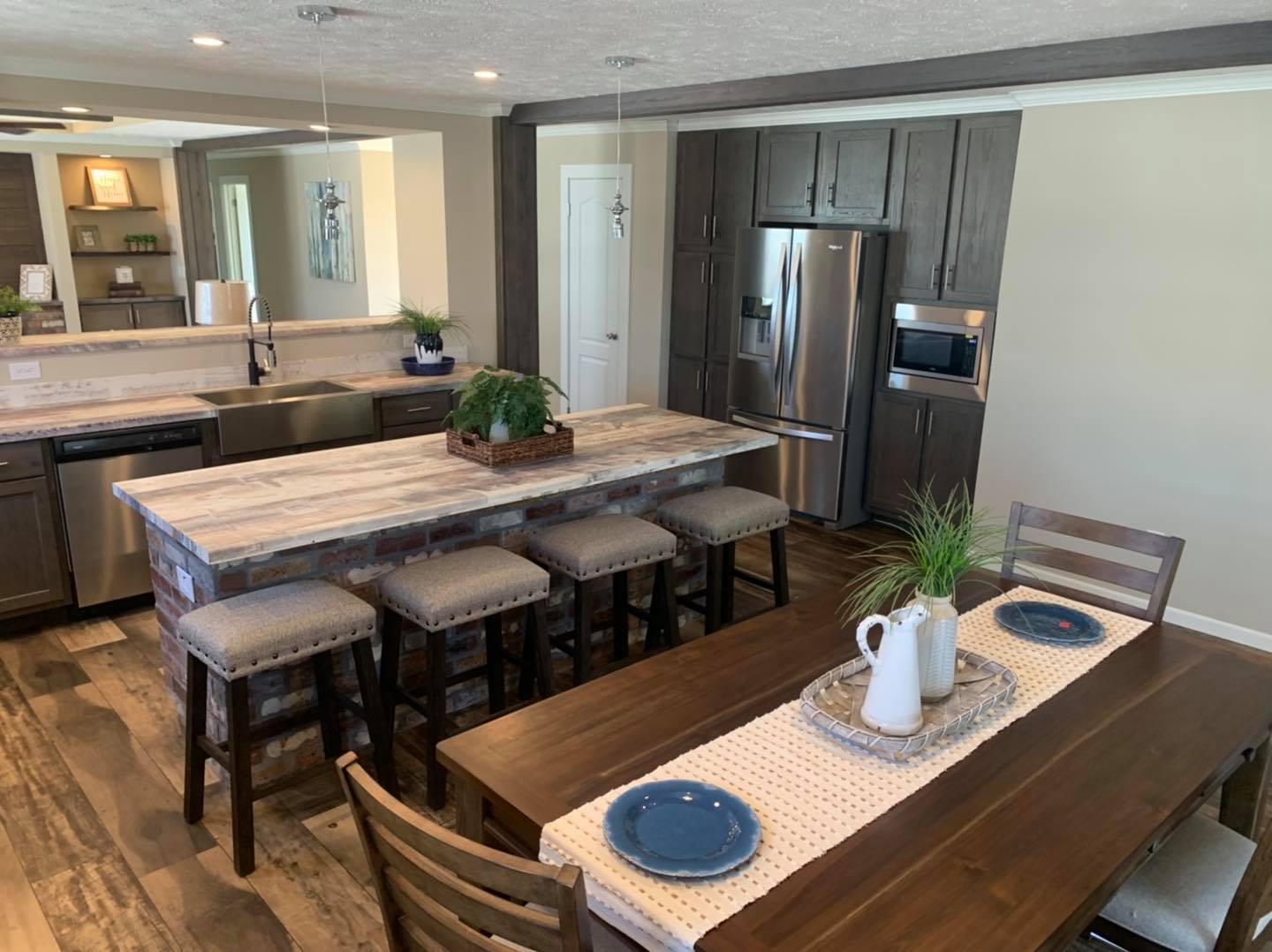 Modern kitchen with a rustic wood dining table, three blue plates, and plants. Brick island with stools, stainless steel fridge, and sleek cabinets.