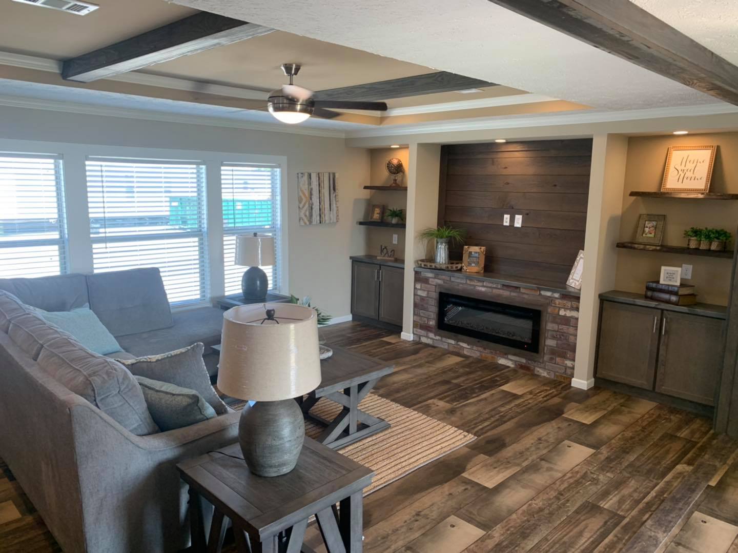 Cozy living room with beige walls and wood-accented ceiling. Features a gray sectional, brick fireplace, and shelves with decor. Bright from large windows.