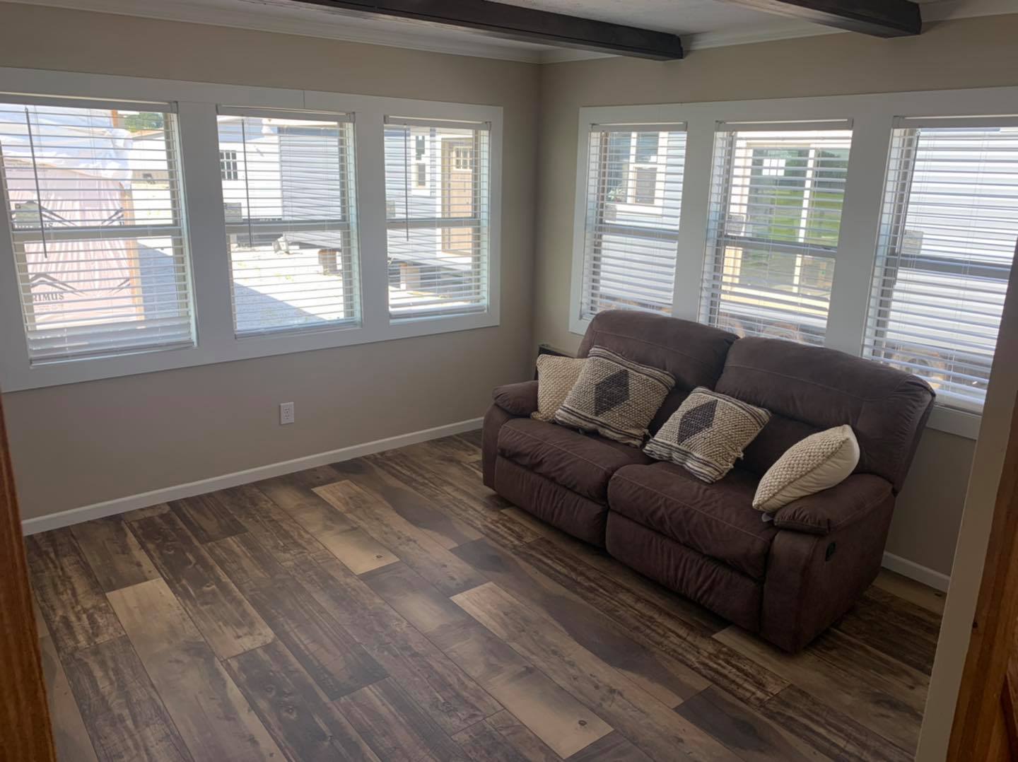 A cozy room with wooden flooring, a brown sofa with patterned cushions, and five large windows letting in natural light. Calm and inviting ambiance.