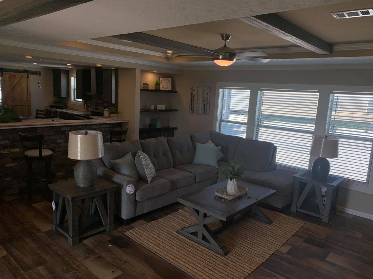 Cozy living room with a gray sectional sofa, wooden coffee table, and side tables with lamps. Large windows with blinds and adjacent open kitchen space.
