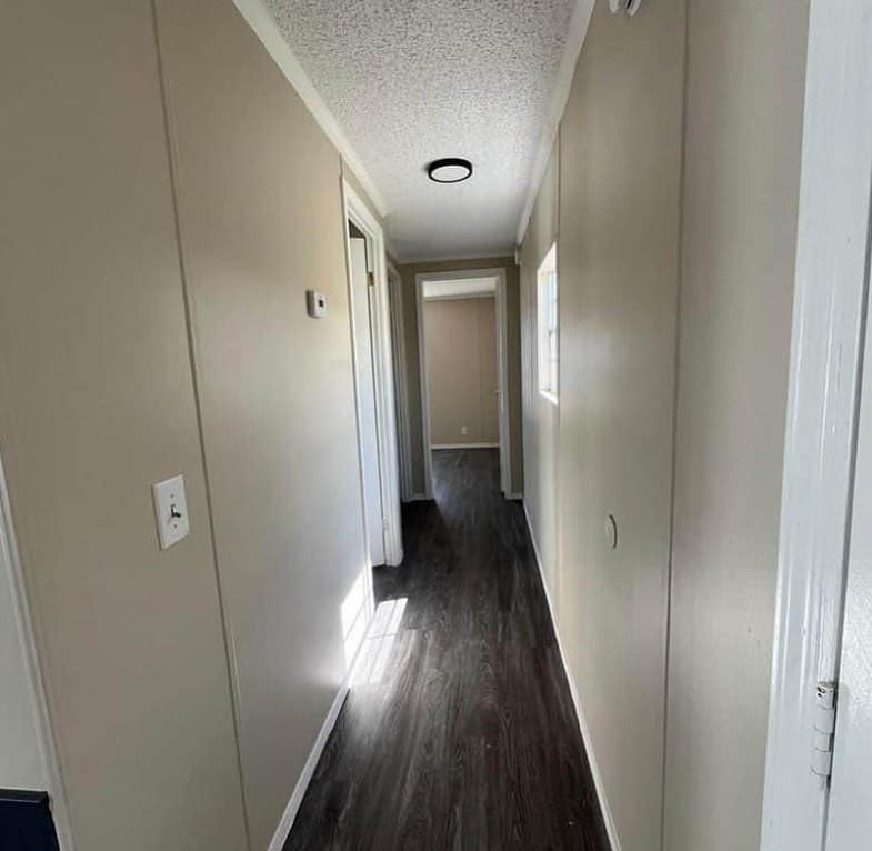 Narrow hallway with beige walls and a dark wooden floor, illuminated by natural light from a window. The space feels clean and minimalist.
