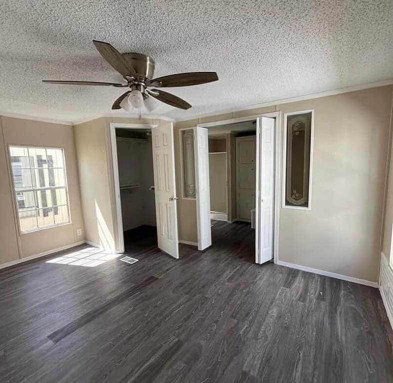 Bright room with beige walls and dark wood flooring. A ceiling fan is centrally located. Two open closets and a window allow light to fill the space.
