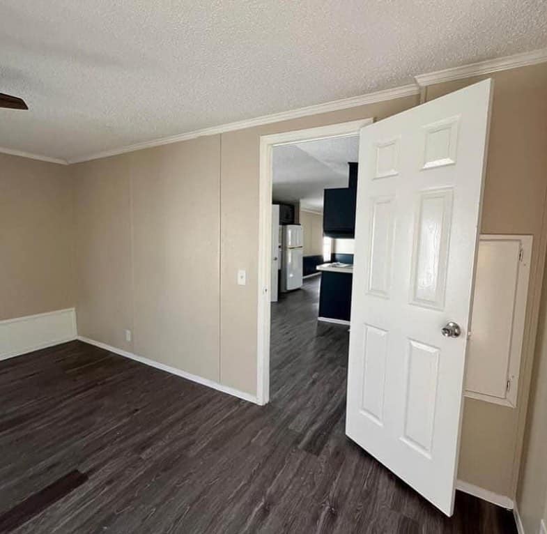 Empty room with beige walls, dark wood flooring, and an open white door. The door leads to a kitchen area with a white fridge and dark cabinets.
