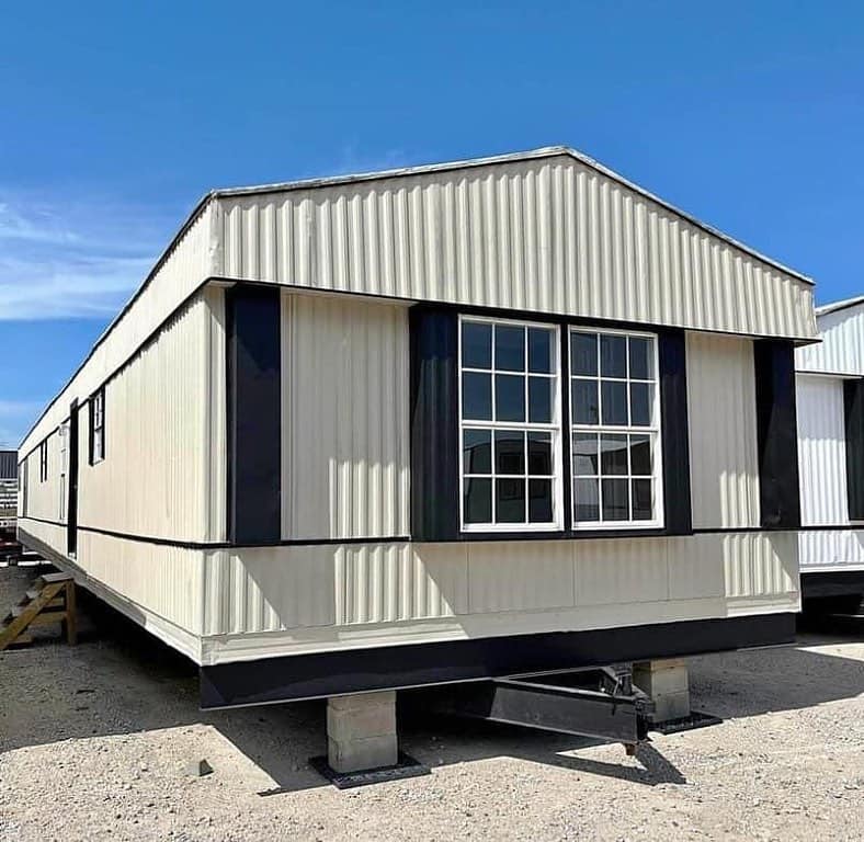 A beige mobile home with a peaked roof stands on concrete blocks against a clear blue sky. It features large windows with black trim.