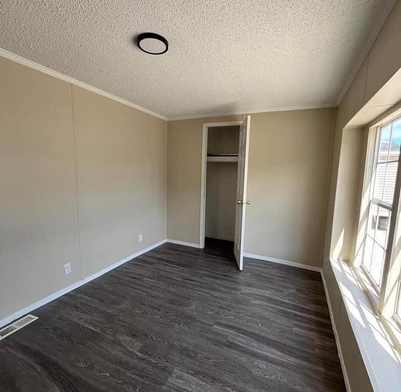 Empty room with beige walls, dark wood floor, and a small closet with an open door. Sunlight streams through a large window on the right.