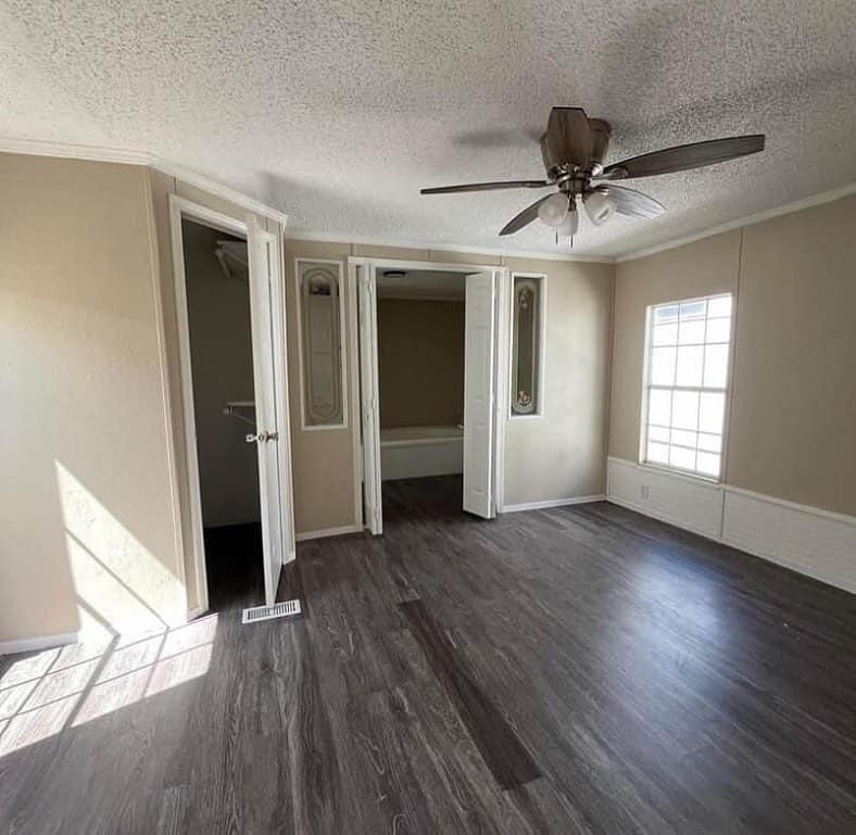 Empty room with light-colored walls and dark wood flooring, featuring a ceiling fan and bright window light. Doorways lead to a bathroom and closet.