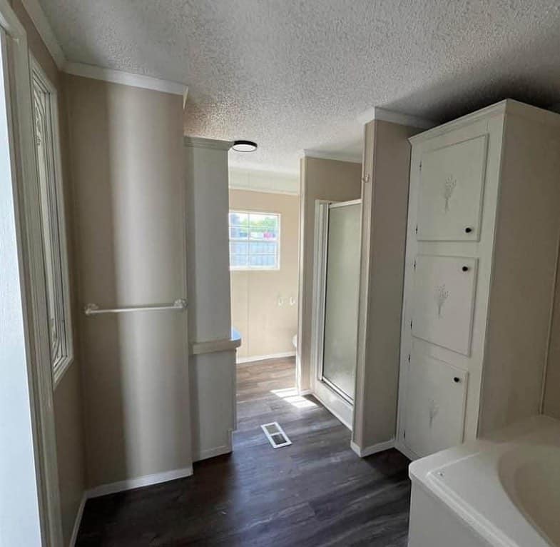 Narrow bathroom with beige walls and dark flooring. Features include a shower with glass door, white cabinets, and a window letting in natural light.