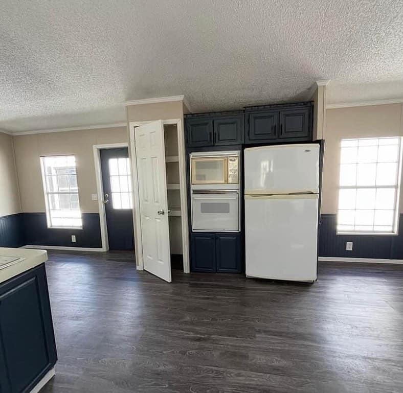 Kitchen with dark wood floor, dark blue lower cabinets, a white fridge and oven. Beige walls, bright light from two windows and a door.