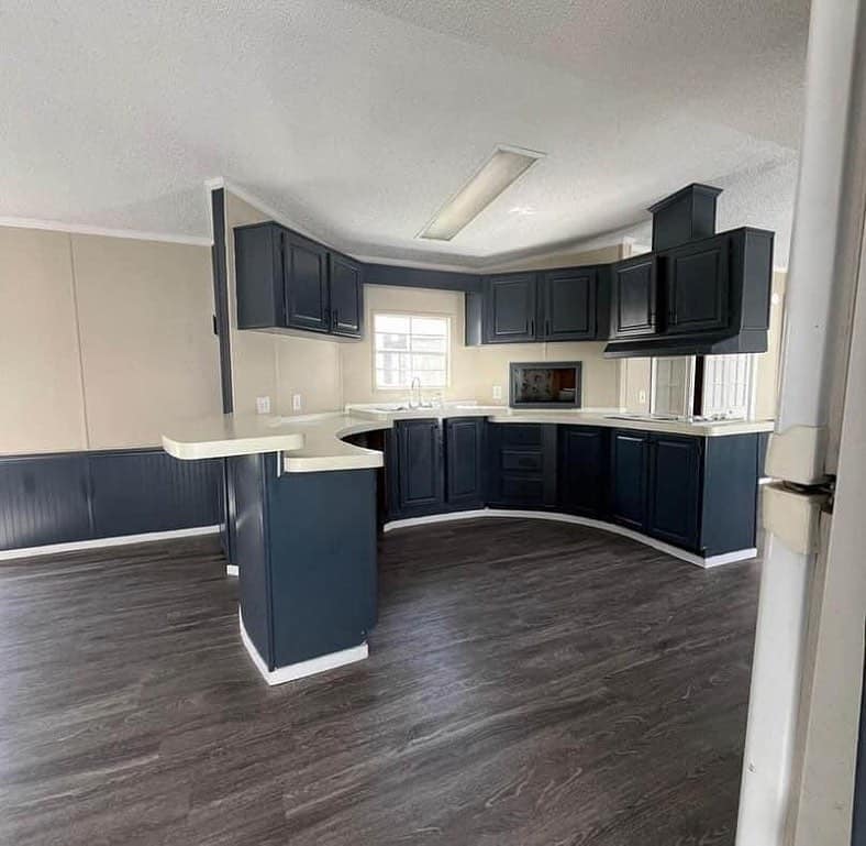 A modern kitchen with dark blue cabinets, white countertops, and dark wood floors. A window provides natural light, creating a clean, open atmosphere.