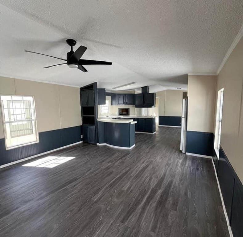 Spacious modern living area with dark wood flooring, off-white and navy walls, a black ceiling fan, and a kitchen with dark cabinets and light countertops.