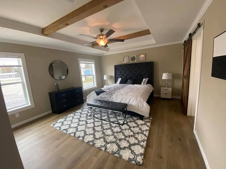 A cozy bedroom with a tufted black headboard, white bedding, and a patterned rug. Wooden beams and a ceiling fan add warmth. Sunlight streams through two windows.