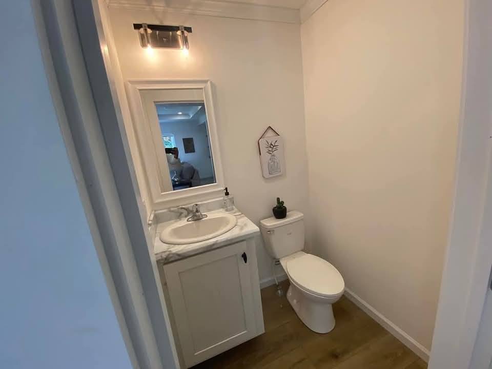 Small bathroom with white walls, featuring a toilet and a vanity with a marble top. A mirror is above the sink, lit by wall sconces. A small plant and decorative art add charm.