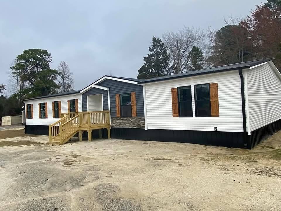 Spacious white and gray mobile home with wooden shutters sits on a sandy lot. It features a small wooden porch, surrounded by trees under a cloudy sky.