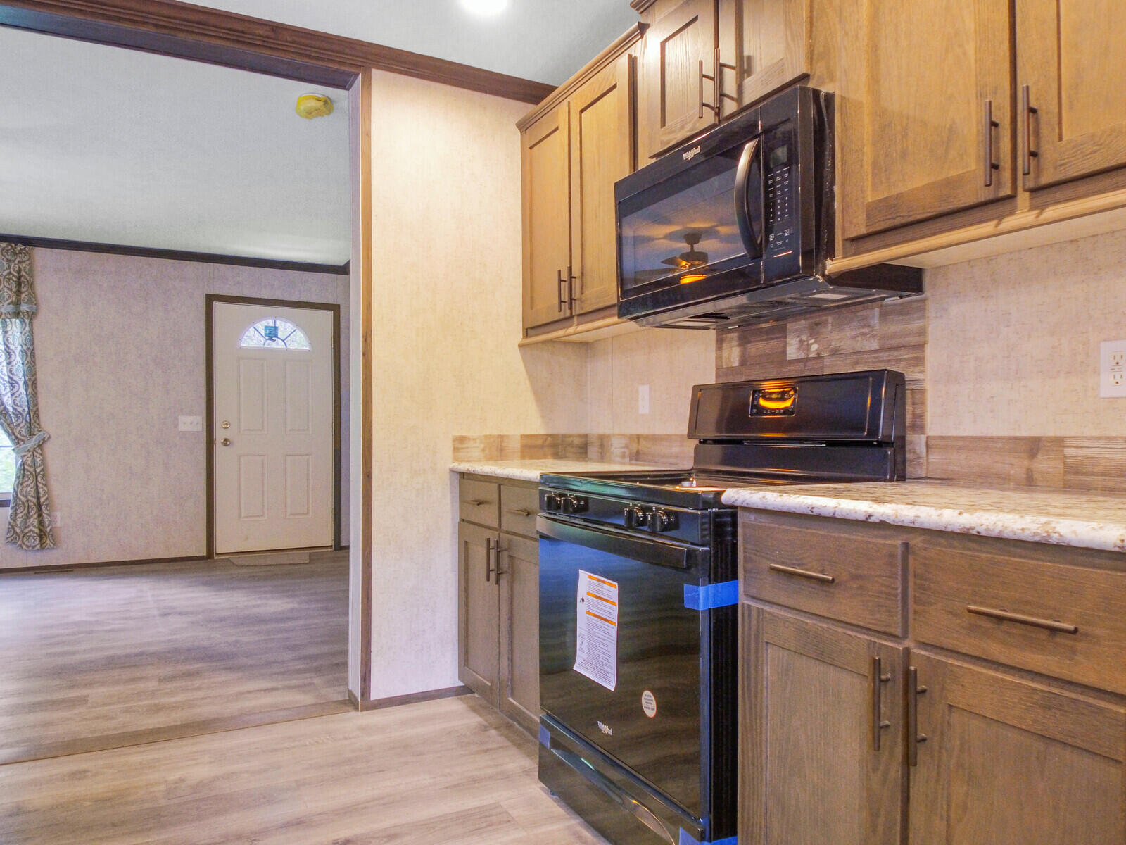 Modern kitchen with wooden cabinets, sleek black appliances, and granite countertops. An adjacent room features light wood flooring and a front door.