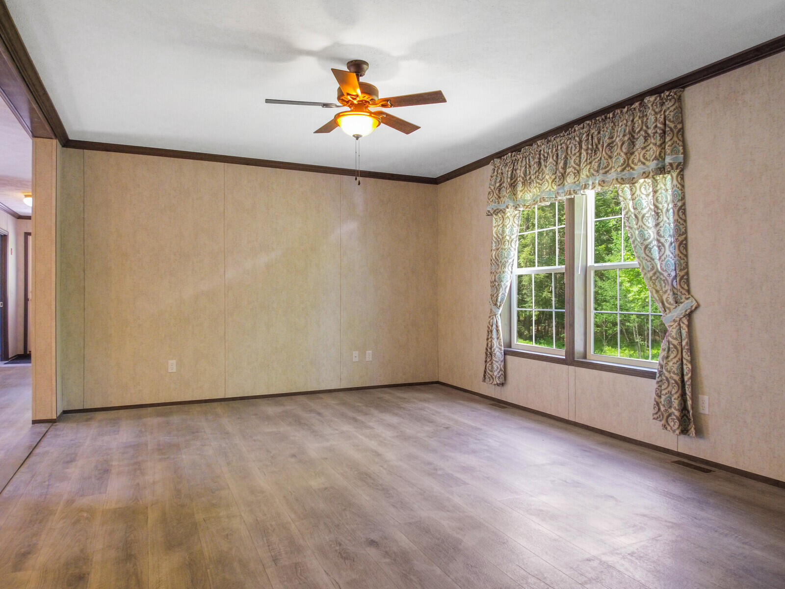 Empty living room with beige walls and wood flooring. A ceiling fan with a warm light hangs above. A large window with patterned curtains overlooks greenery.