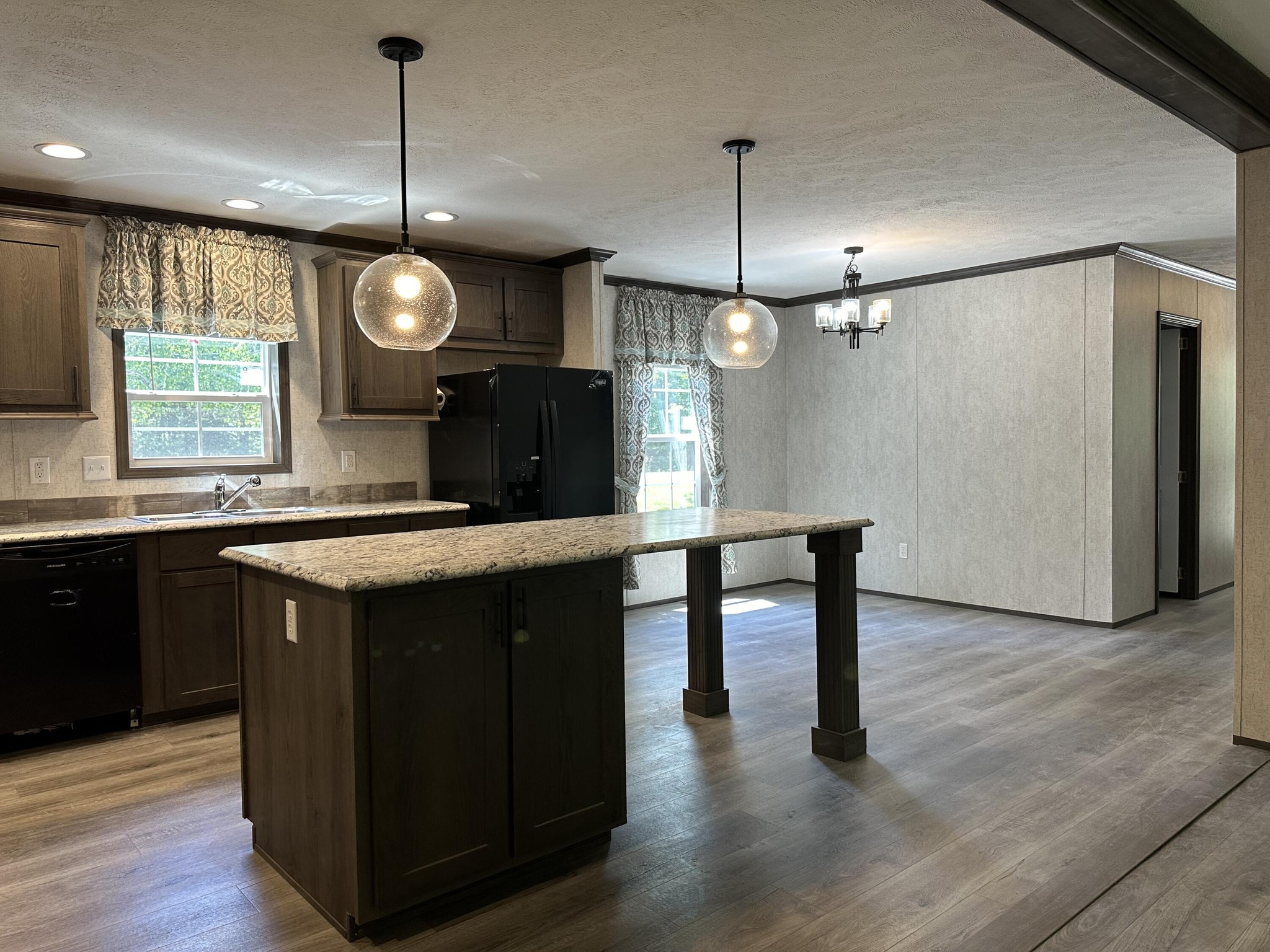 Modern kitchen with dark wood cabinets, granite countertops, and two pendant lights hanging above a central island. Large windows with patterned curtains brighten the space's neutral tones.
