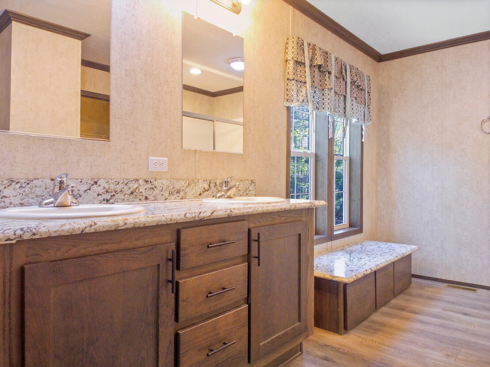 Elegant bathroom with dual sinks on a granite countertop, wooden cabinetry, large window with patterned curtains, and a cozy bench. Warm, inviting tone.
