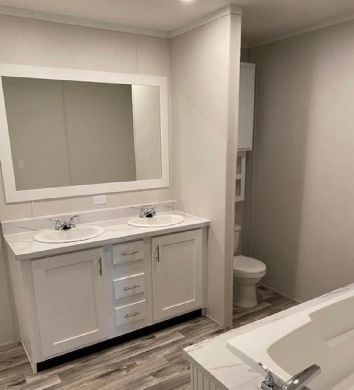 A modern bathroom features a double sink vanity with a large mirror, white cabinetry, and a marble countertop. A bathtub and toilet are visible, with wood-like flooring enhancing the warm, clean aesthetic.