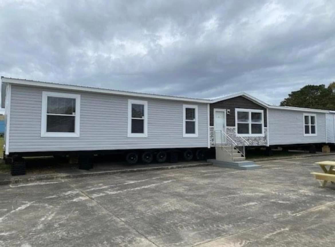 A gray double-wide trailer home with white trim and multiple windows sits elevated on wheels. A small set of stairs leads to the front door. Cloudy sky above.