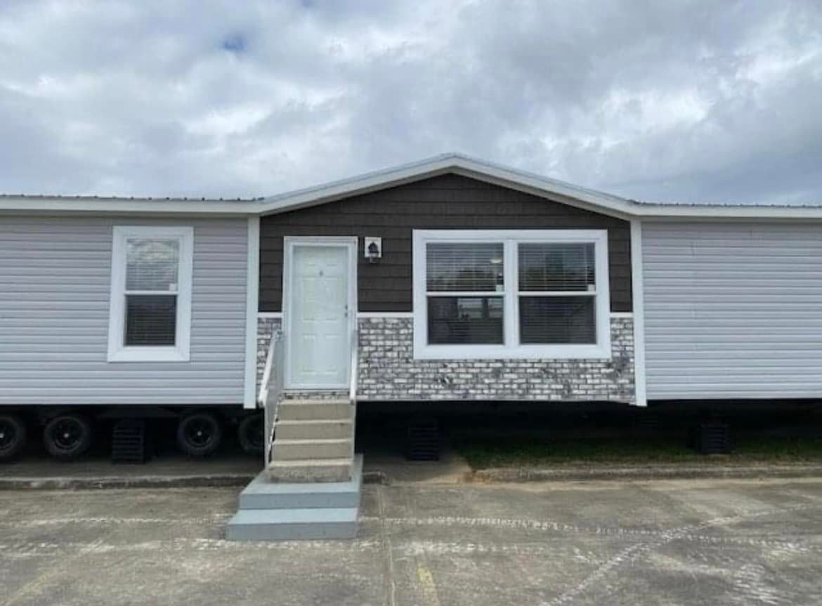 A single-story modular home with white siding and a brick accent. It features a centered door with steps, two windows, and a cloudy sky above.