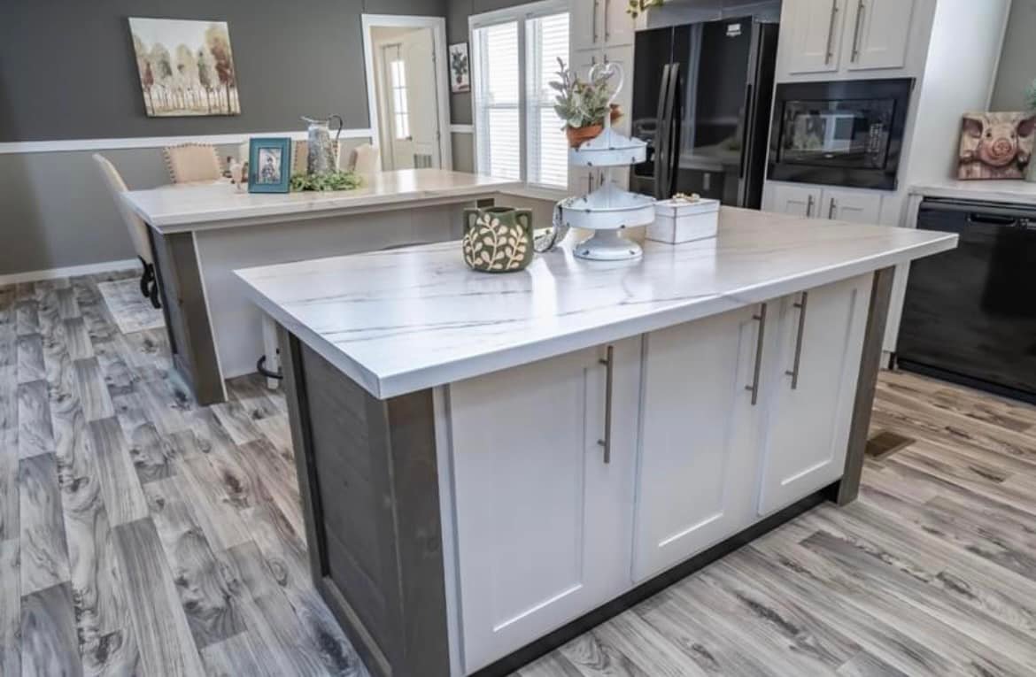 A modern kitchen with two white marble islands, grey cabinets, wood floors, and black appliances. Decor includes plants and white tiered trays.