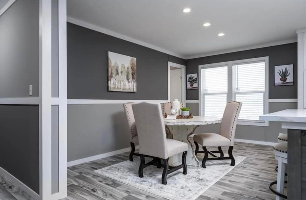 Dining room with a round table and four beige upholstered chairs on a patterned rug. Gray walls, landscape painting, and two windows create a cozy atmosphere.