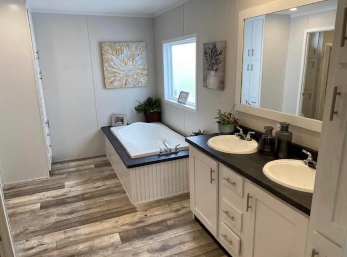 A modern bathroom with light wood flooring features a white bathtub, dual sinks, and dark countertops. Natural light floods a serene, tidy space. 