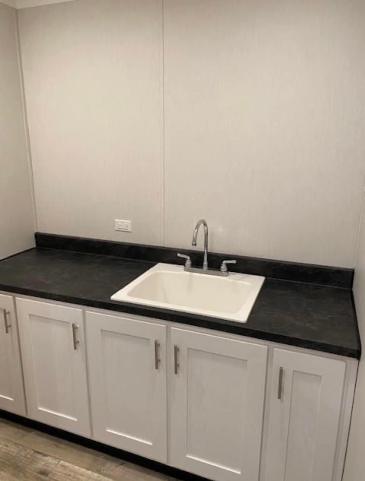 A minimalist kitchen with a sleek black countertop and white cabinets. A white sink with a modern faucet is centered, against a plain light wall.