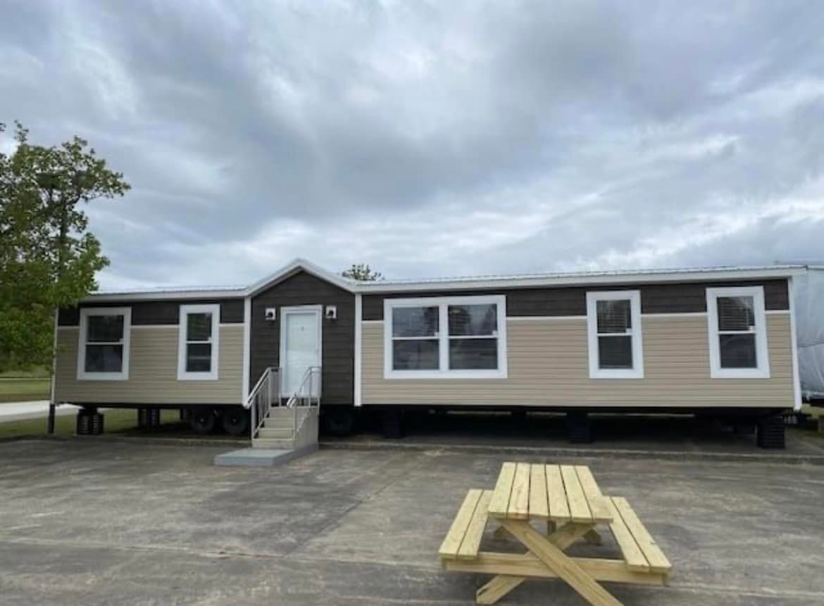 A beige and brown modular home sits on a concrete lot under a cloudy sky. Steps lead to the front door centered between windows. A wooden picnic table is in the foreground.