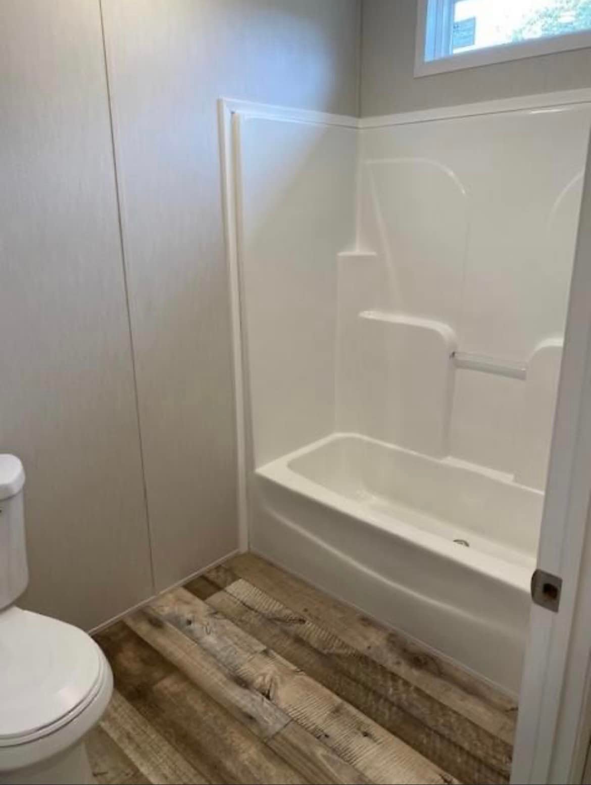 Minimalist bathroom with white bathtub and shower combo, wooden floor, and neutral walls. A window lets in natural light, enhancing the clean look.
