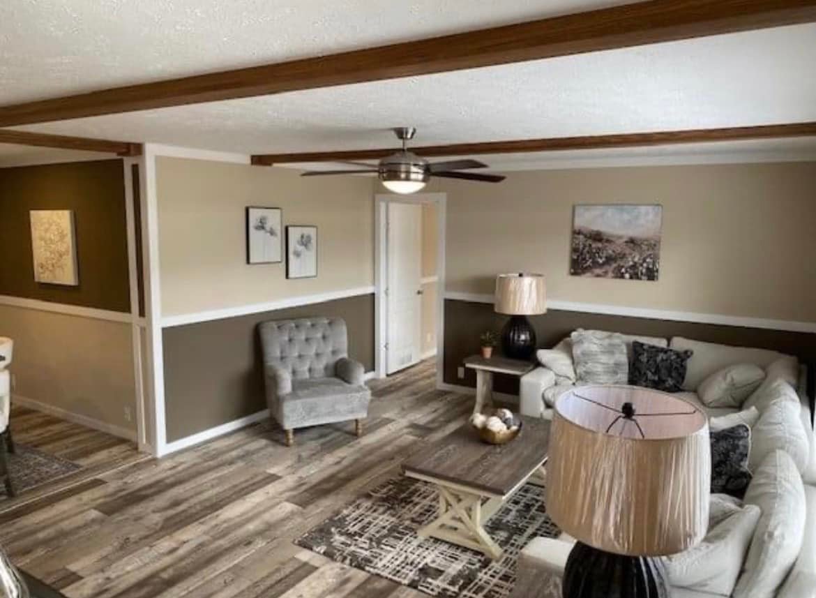 Spacious living room with a light beige and brown color scheme. Features a gray armchair, sectional sofa, ceiling fan, and rustic wood floors. Cozy and inviting.