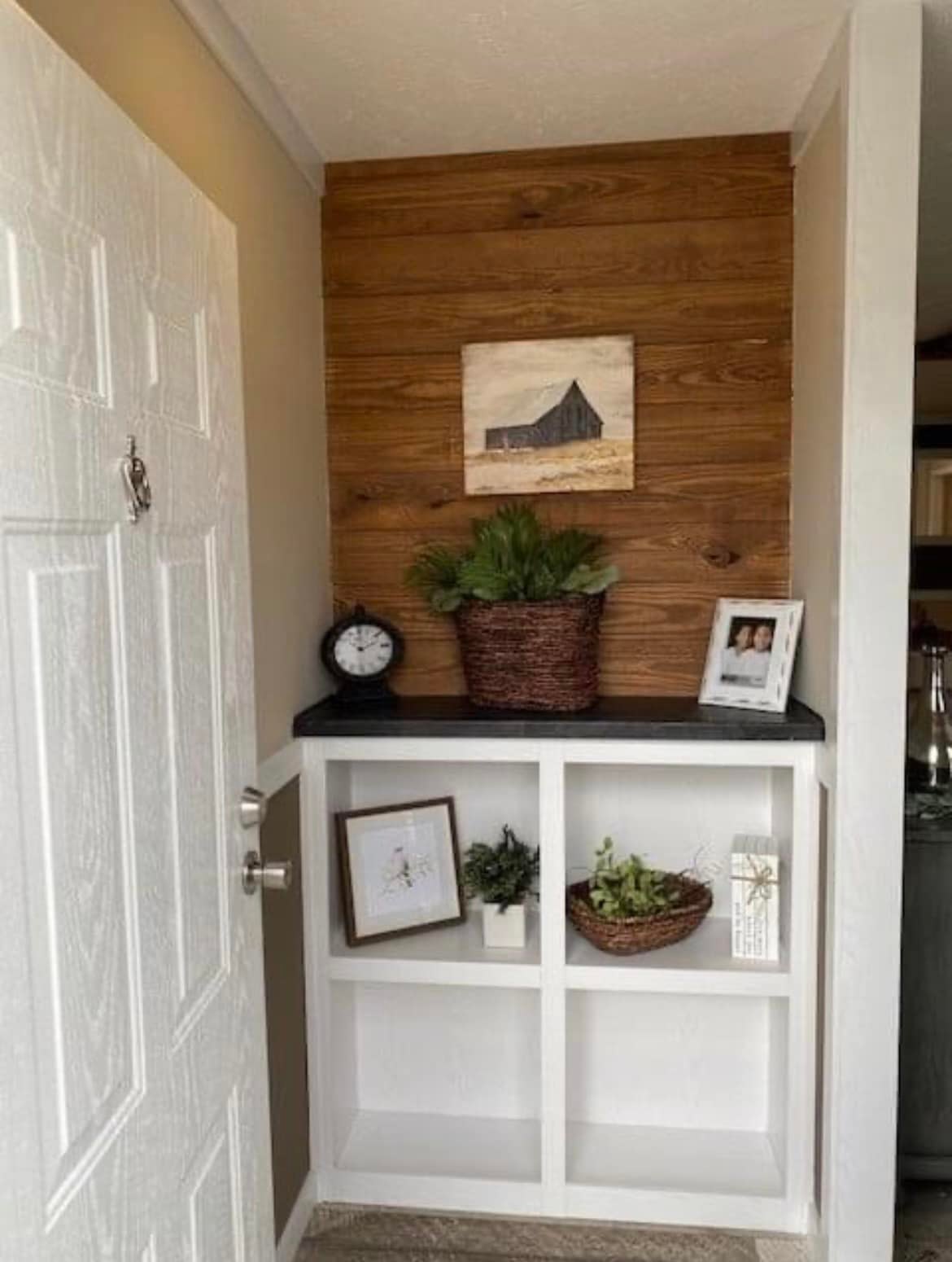 Cozy entryway with wooden accent wall, white shelves, and décor. Features a clock, potted plants, artwork, and a framed photo, creating a welcoming ambiance.