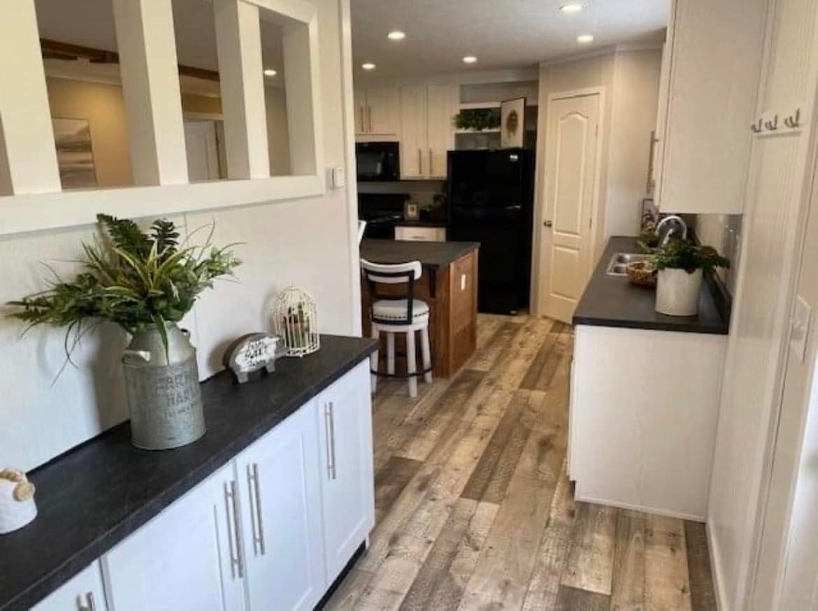 Modern kitchen with white cabinets, black countertops, and wood flooring. Features potted plants, decorative items, and a small island with stools. Cozy and inviting.