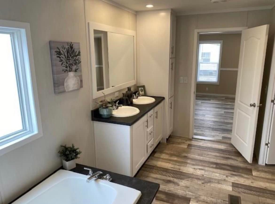 Modern bathroom with light wood flooring, a double sink vanity with dark countertop, wall art, and a window. Open door leads to a bright room.