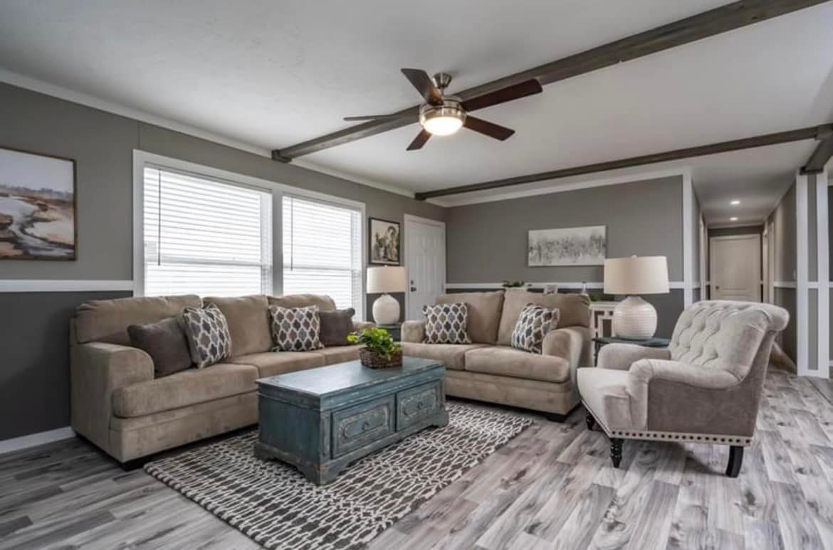 Modern living room with gray walls, beige sofas, patterned cushions, blue coffee table, and armchair. Ceiling fan and natural light enhance coziness.