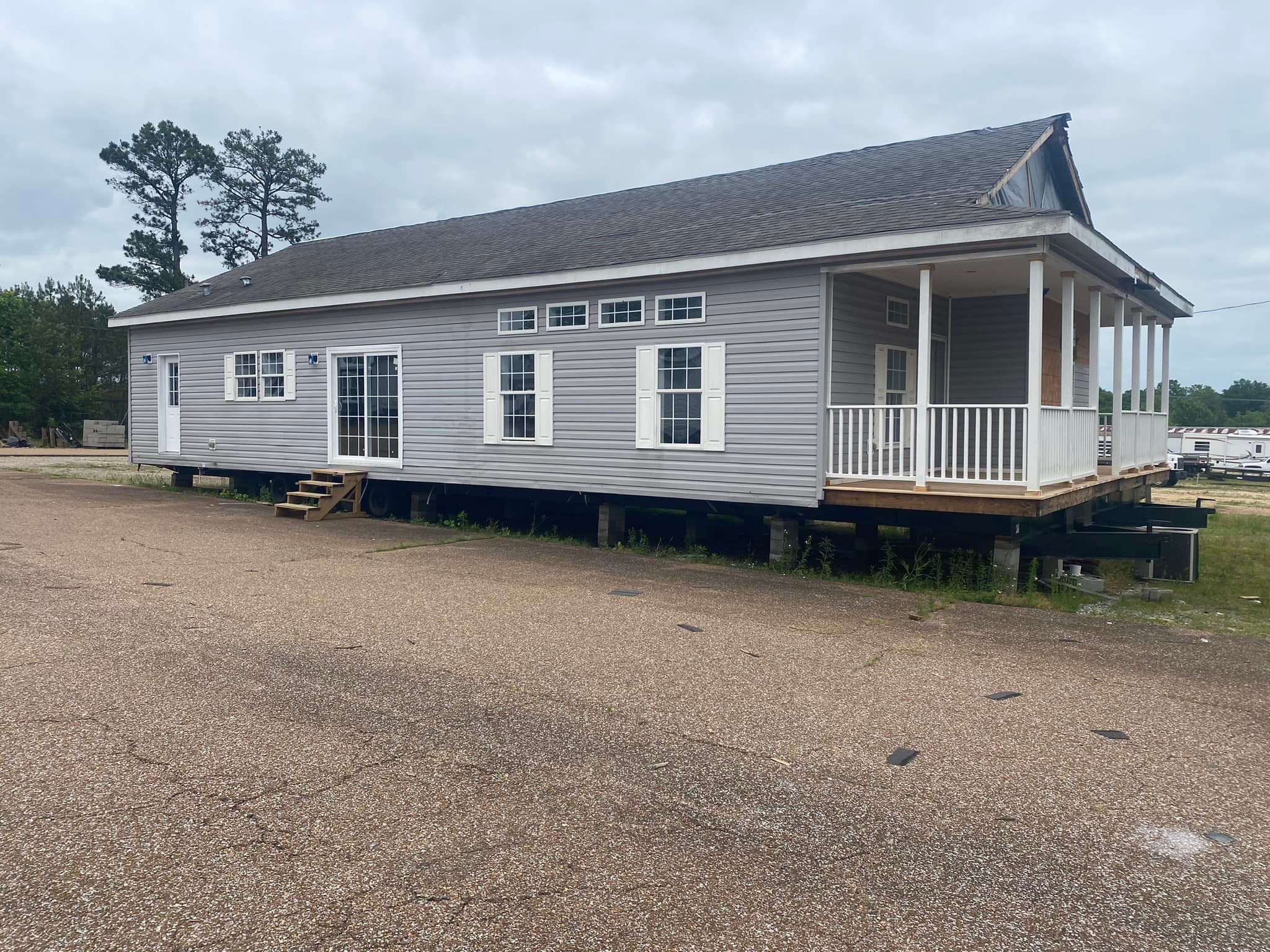 A gray modular home on blocks, featuring a wraparound porch with white railings. Overcast sky and trees are visible in the background.