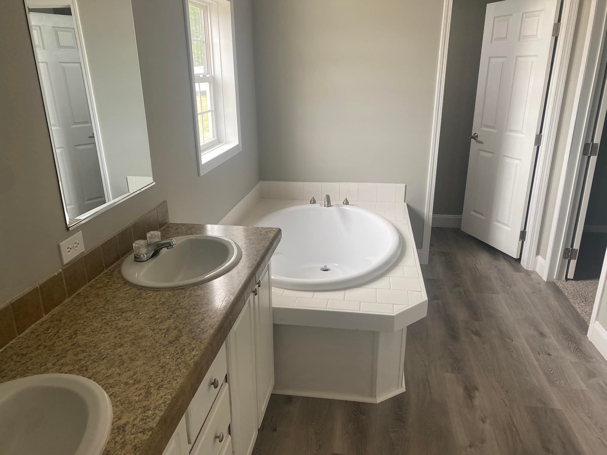 Bathroom with gray walls, featuring a white drop-in bathtub beside a wooden vanity with a beige countertop and single sink. Light streams through a window.