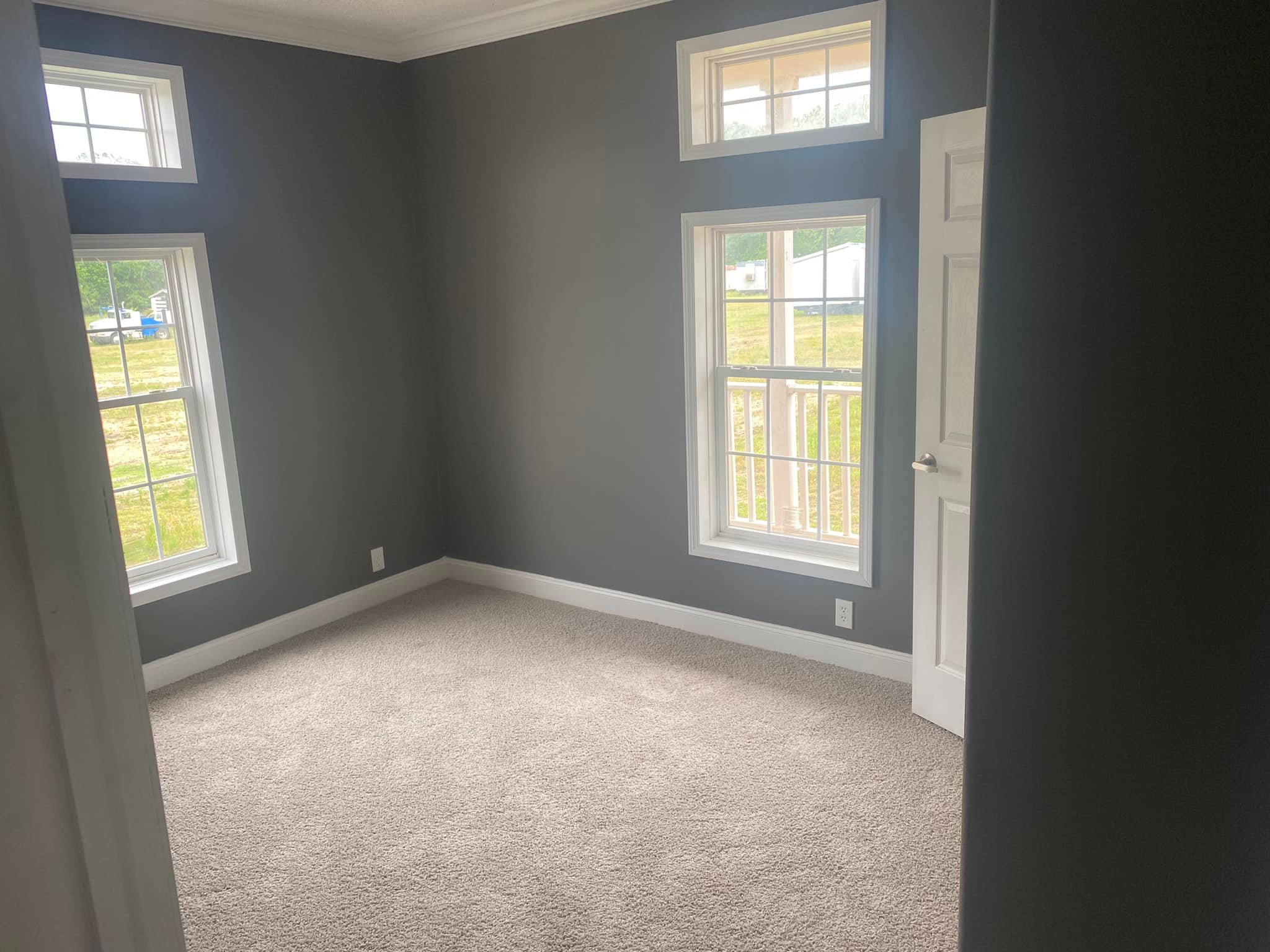 A small, empty room with gray walls and beige carpet. Two tall windows let in natural light, offering a view of grass outside. The mood is calm and minimalistic.
