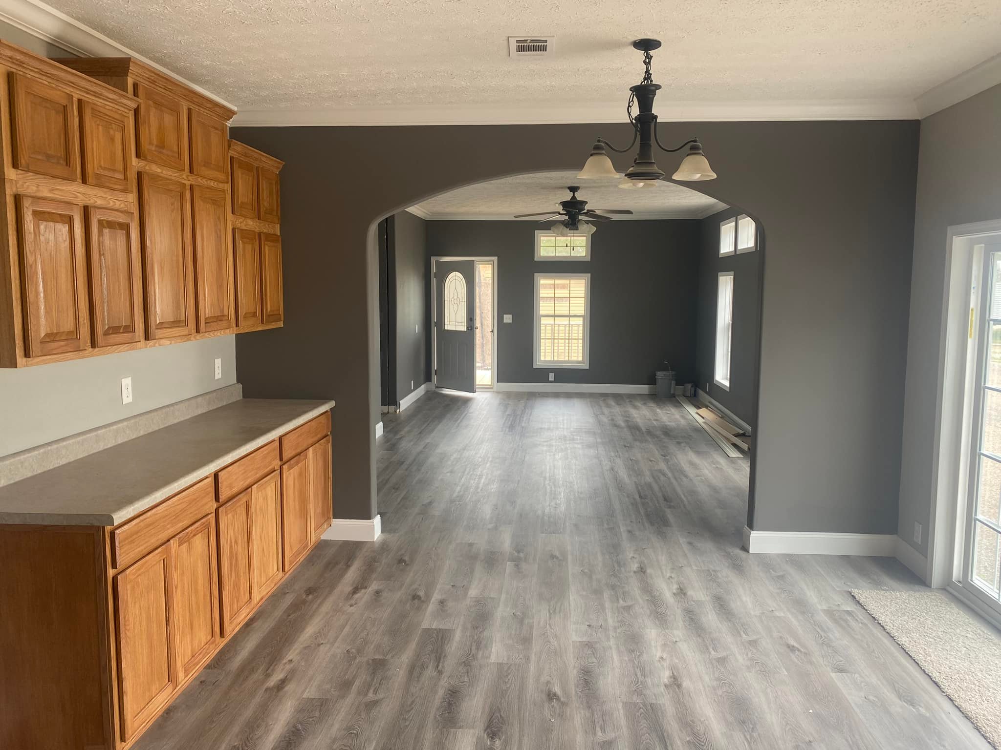 Spacious room with gray walls and hardwood floors, featuring wooden cabinets on the left. Arched doorway leads to another room with a windowed door. Neutral tones and a calm ambiance.