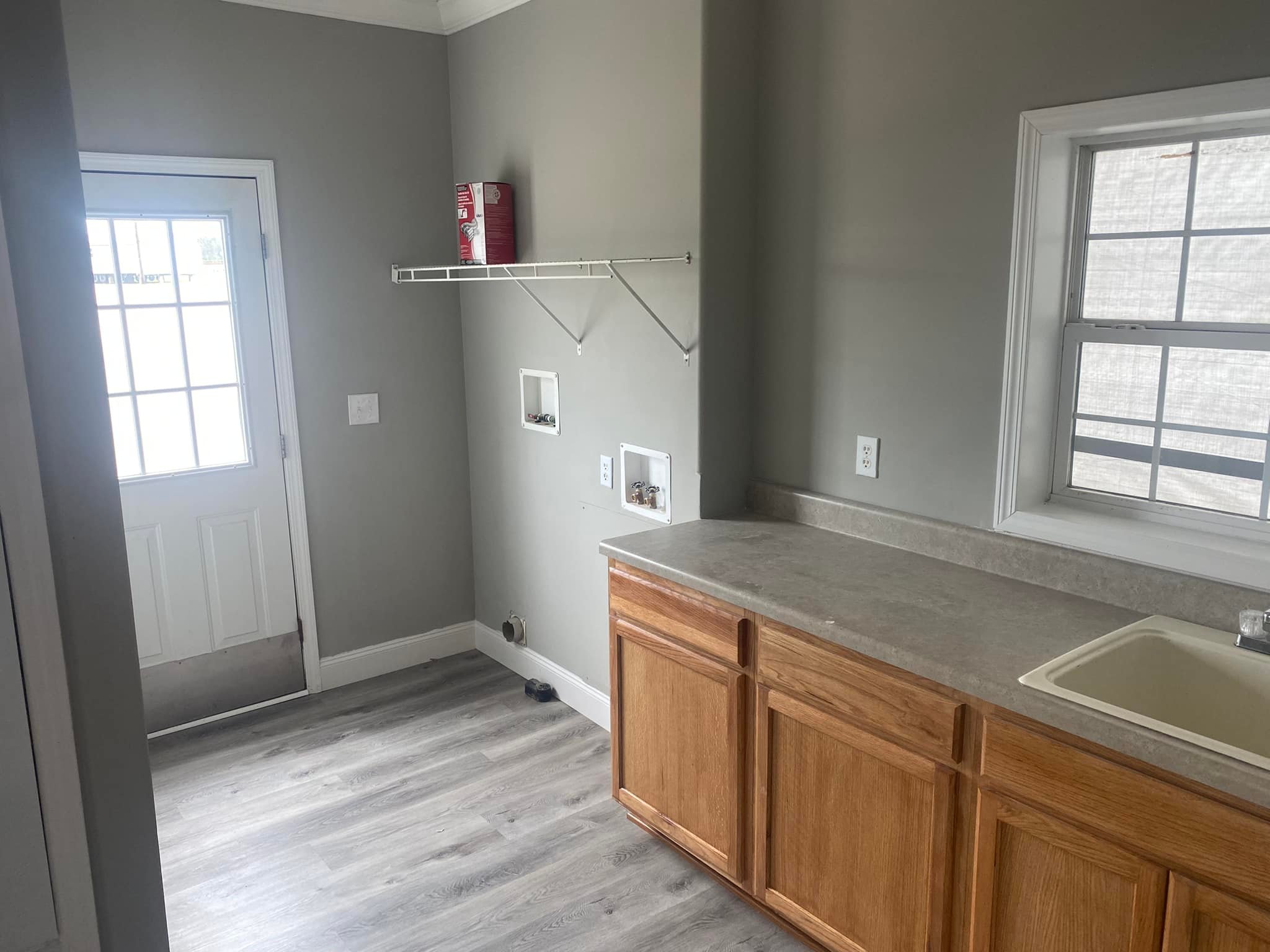 Laundry room with light gray walls and laminate wood flooring. Features a washing machine hookup, a wooden cabinet with a sink, and a window.