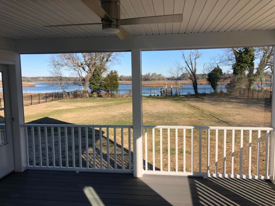 Covered porch with a ceiling fan overlooking a grassy yard and a peaceful lake with trees. The scene conveys a serene and tranquil atmosphere.