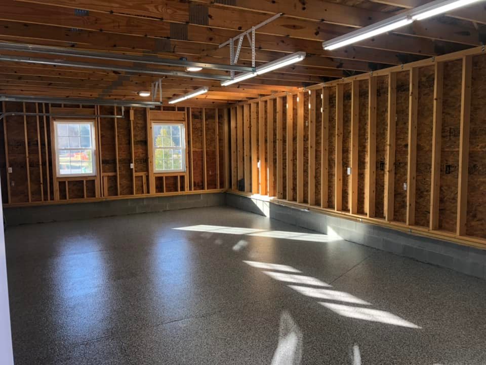 Spacious unfinished room with exposed wooden framing and two windows. Smooth, speckled concrete floor, lit by bright fluorescent ceiling lights.