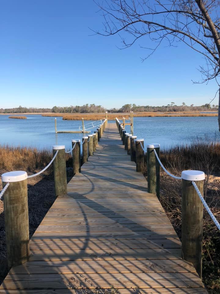 Wooden dock extending over a calm, blue lake with surrounding marsh grass. Clear sky and tree branches frame the serene, peaceful scene.