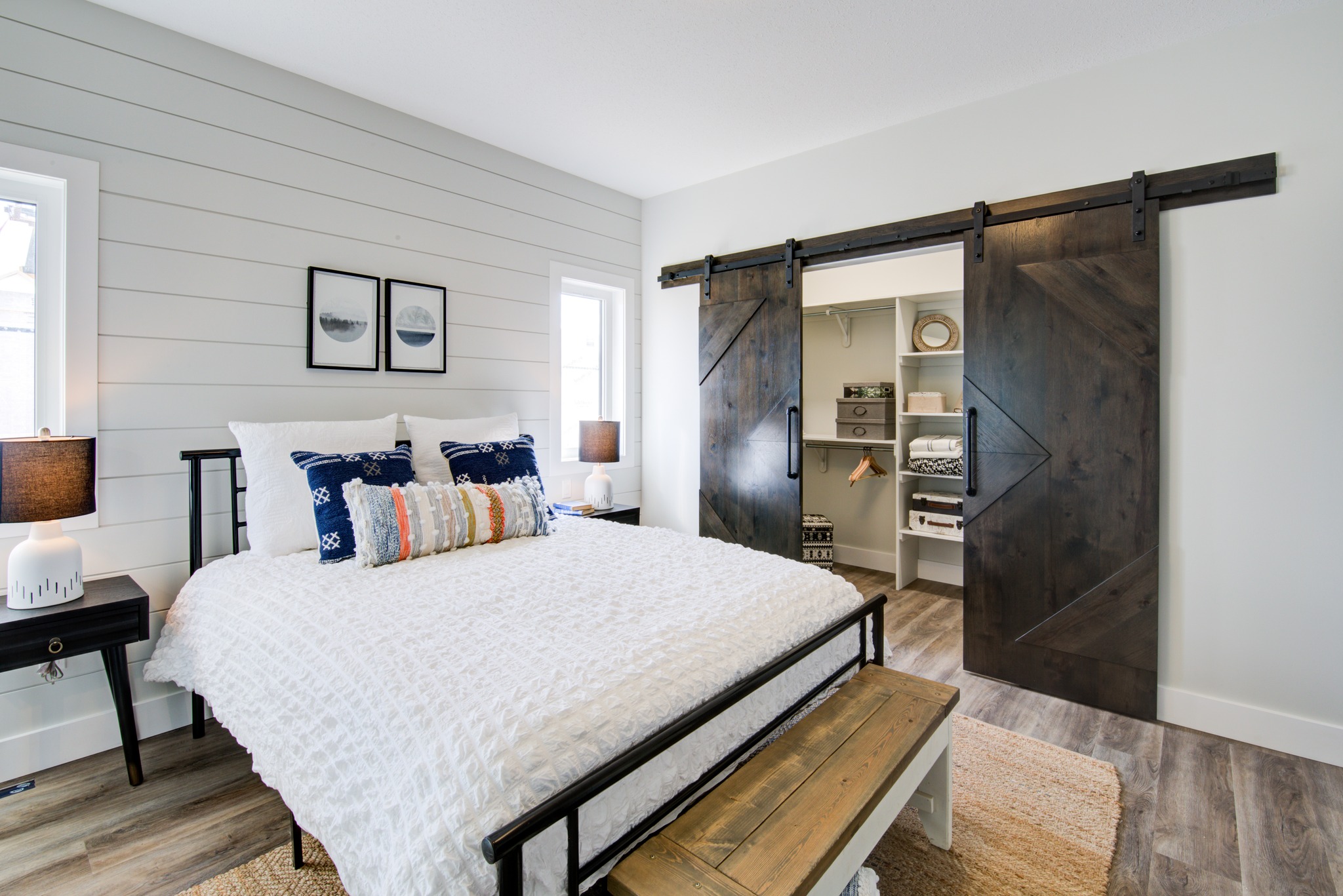 Bright bedroom with a white bed, decorative pillows, and dark wood sliding barn doors revealing a closet. Wooden bench adds rustic charm.