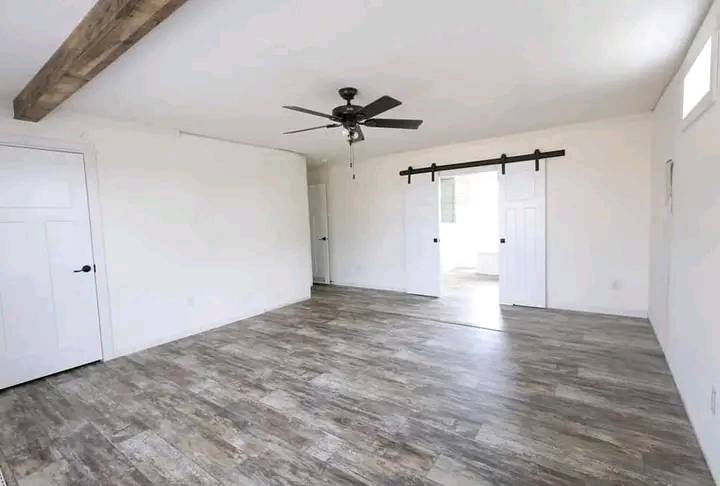 Spacious empty room with white walls, rustic wood flooring, a ceiling fan, exposed wooden beam, and sliding barn door. Bright, airy, and modern feel.