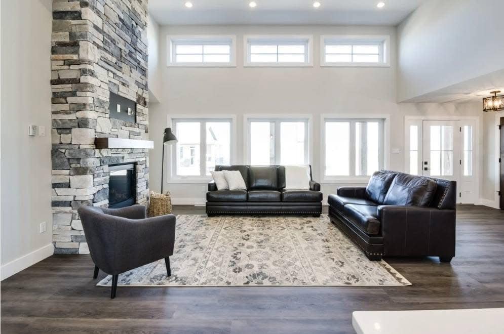 Spacious living room with high ceilings, large windows, and a stone fireplace. Features dark leather sofas, a gray armchair, and a patterned rug.