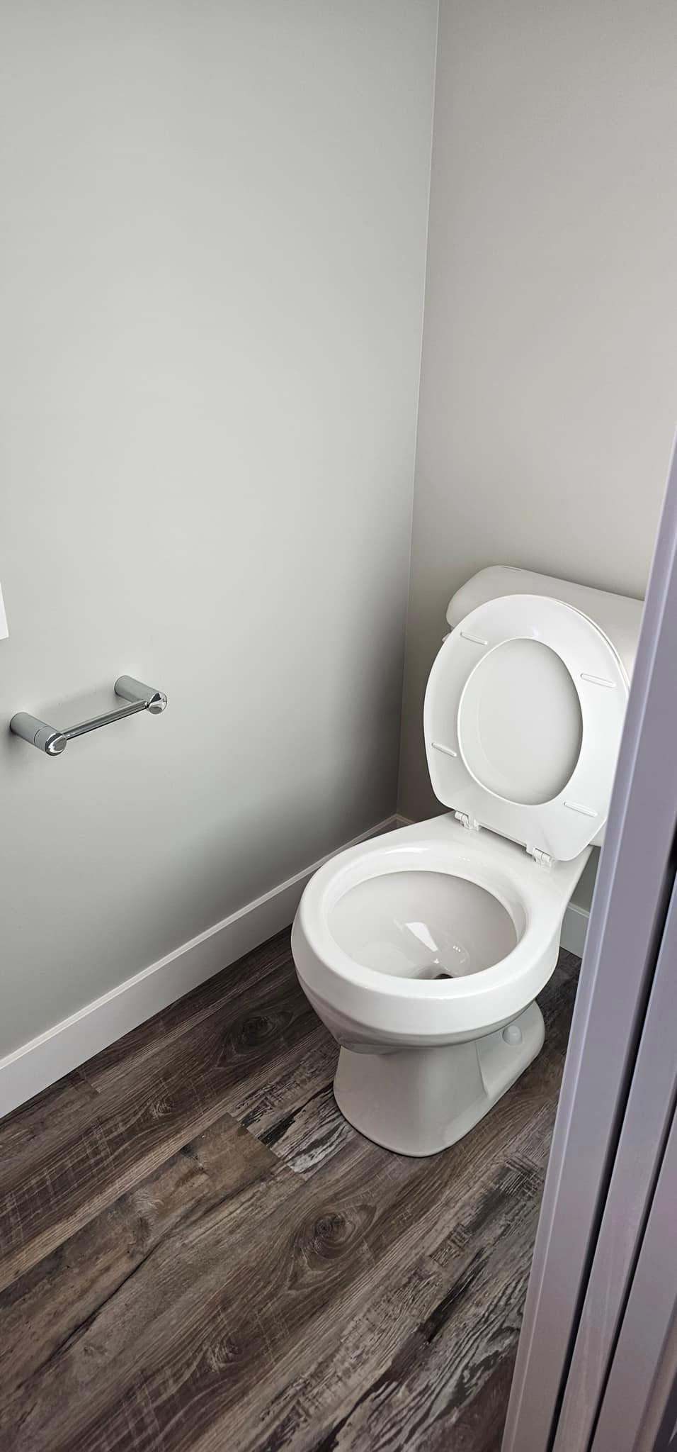A clean, white toilet with an open seat is in a compact bathroom with gray walls and wood-patterned flooring. An empty toilet paper holder is fixed to the wall.