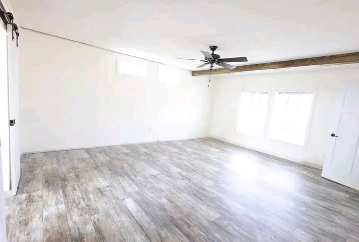 Spacious empty room with light wood flooring, white walls, and a dark ceiling fan. Two large windows allow ample natural light, creating an airy feel.