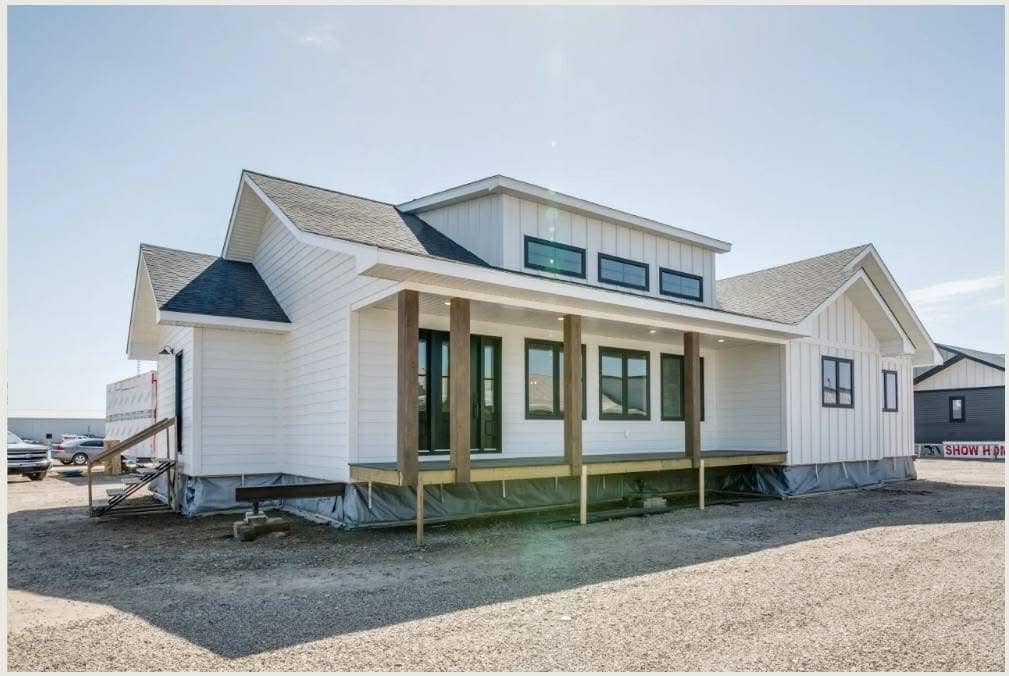 Modern white prefab home with large windows and a small porch, set on a gravel lot under a clear blue sky, conveying a sense of simplicity and innovation.